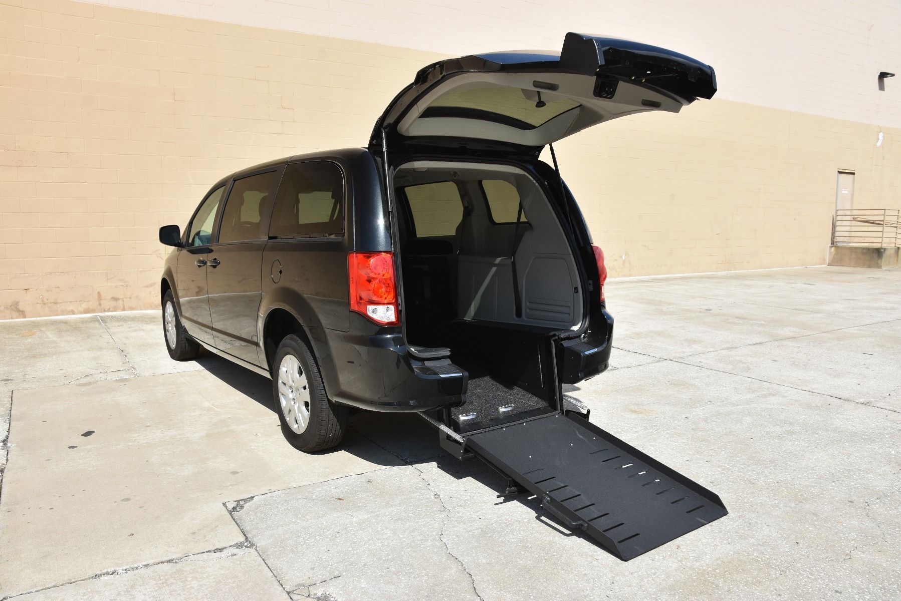 A wheelchair accessible Dodge Grand Caravan minivan in Black with a wheelchair ramp deployed from the open rear hatch.