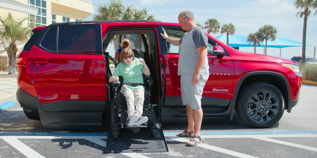 A red wheelchair accessible SUV with ramp deployed from passenger side rear door, with person in wheelchair coming down ramp.