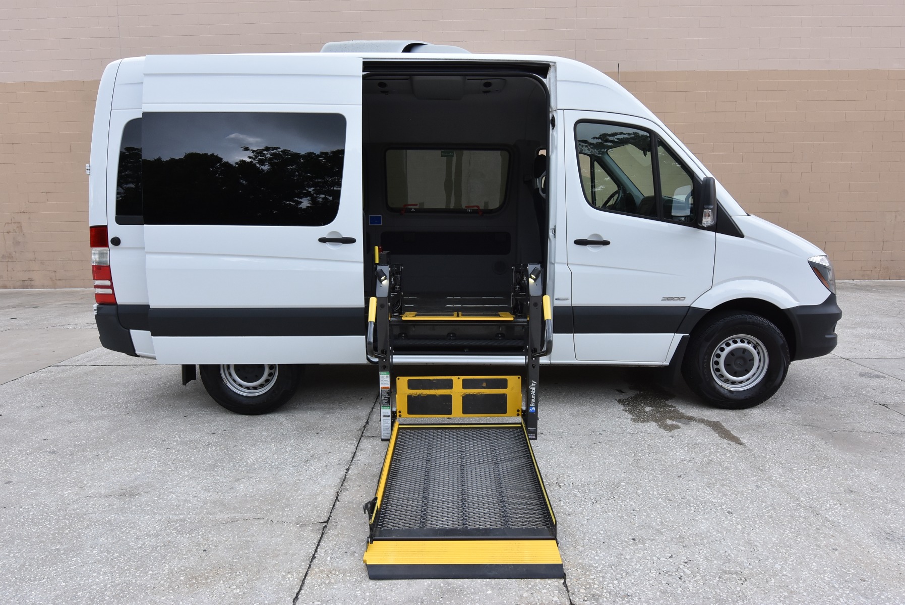 White Mercedes Sprinter full size wheelchair van with lift deployed from open passenger side sliding door.