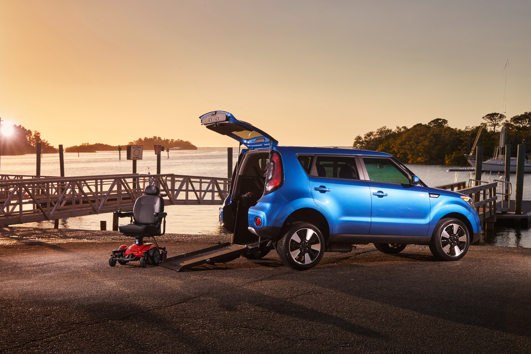 A blue Kia Soul converted for wheelchair access by Ryno Mobility, parked near a dock with ramp deployed from open rear hatch, and a power chair next to the ramp. Sunset in the background.
