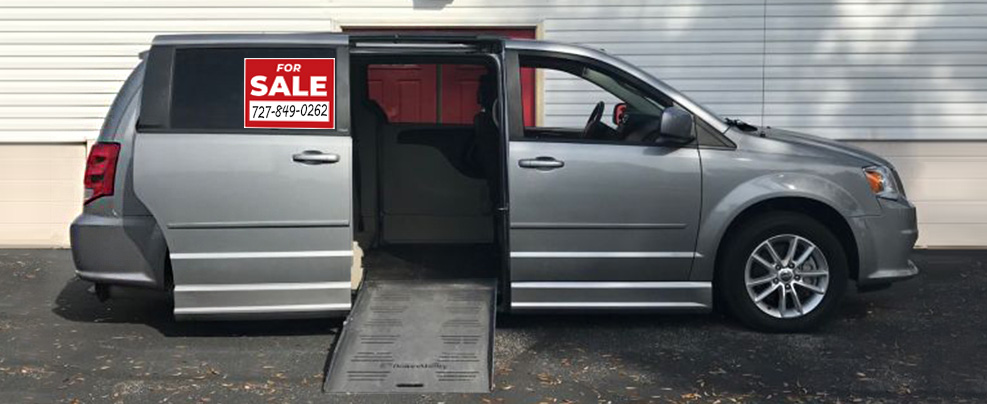 A silver wheelchair accessible minivan is parked on front of a white building. The passenger sliding door is open and there is a ramp deployed for a wheelchair to use.