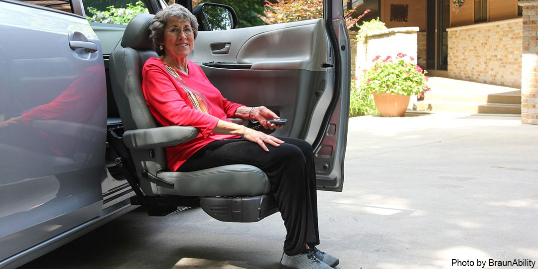 A woman in a pink shirt is sitting on the passenger seat of a vehicle and holding a remote. The turny seat is lowered to the ground via its modification for ease of entry.