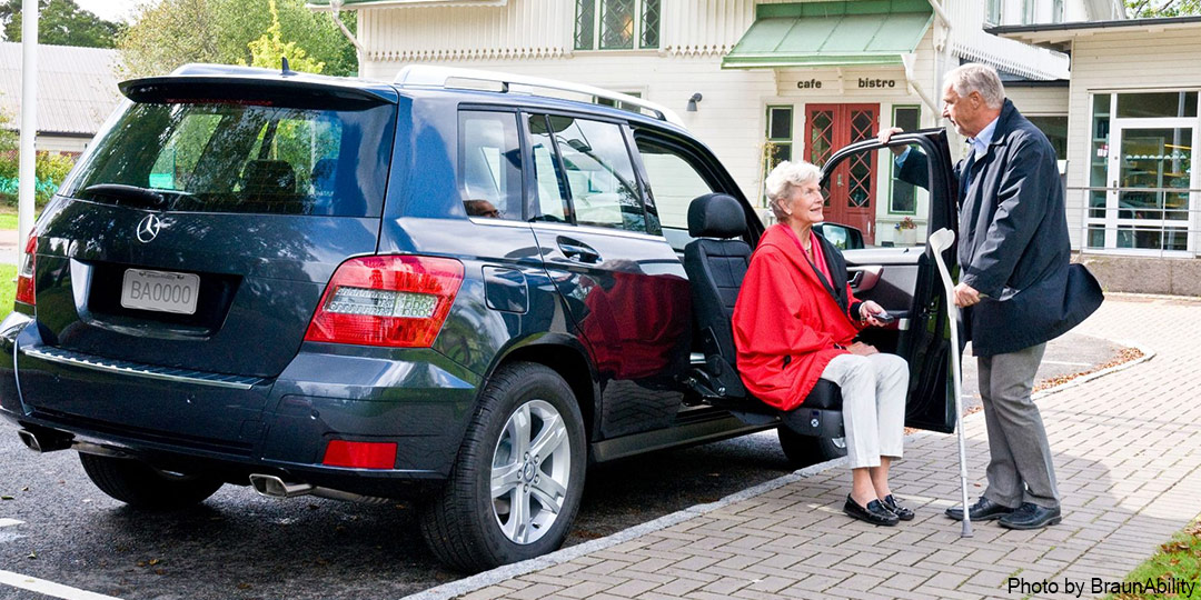 In front of a care, a man is standing on the passenger side of a blue SUV. The passenger door is open, and a woman is sitting on the seat, which has turned and lowered towards the ground to make it easier for her to disembark.