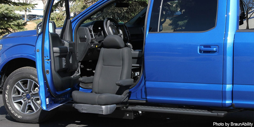 A blue Ford F-150 pickup truck is seen with the driver's door open and the unoccupied seat lowerd towards the ground for ease of entry.